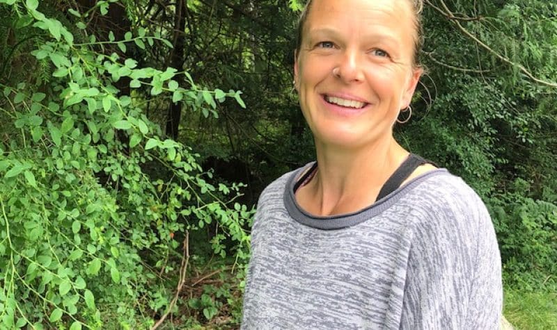 A smiling blonde haired woman in a grey shirt stands in front of a forest.
