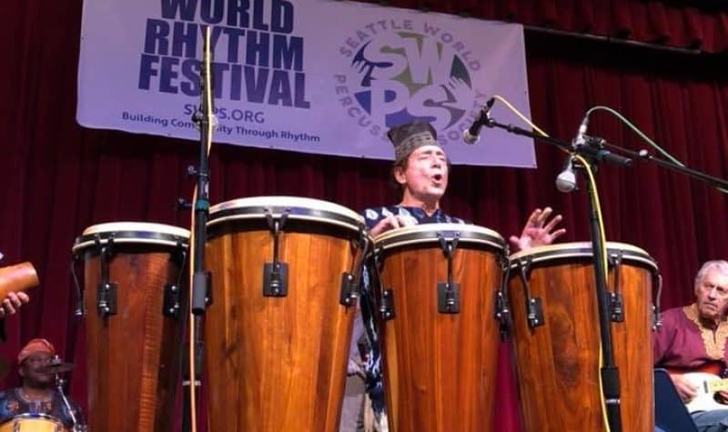 A man on a stage plays a set of five tall hand drums.